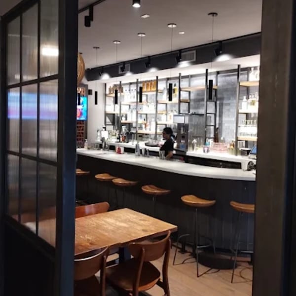 A bartender works behind a well-stocked bar with stools, seen from a dining area with tables.