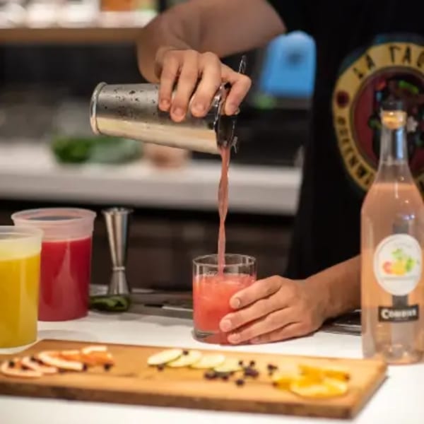 Bartender pouring a red drink from a shaker into a glass, with fruit garnishes and juices on the counter.
