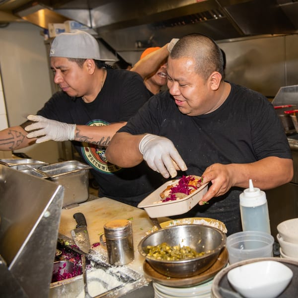 Three La Taqueria chefs in a busy kitchen preparing Mexican food with fresh ingredients.