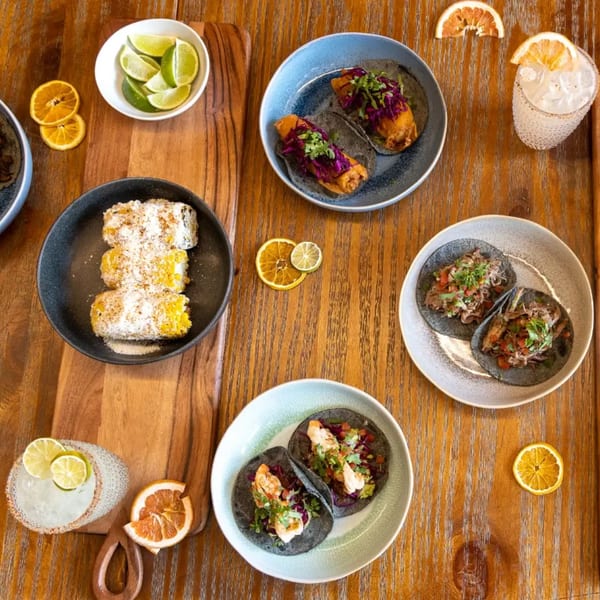 Overhead view of various Mexican dishes including tacos, elote, and margaritas on a wooden table.