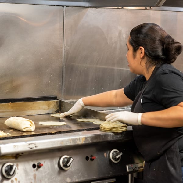 A cook in white gloves prepares tortillas and a burrito on a large griddle at La Taqueria.