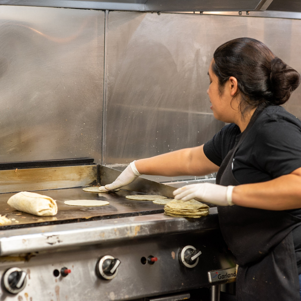 A cook in white gloves prepares tortillas and a burrito on a large griddle at La Taqueria.