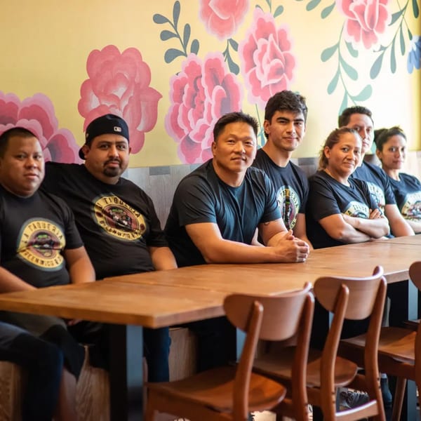 La Taqueria staff in black t-shirts sitting at a long wooden table with a floral wall.