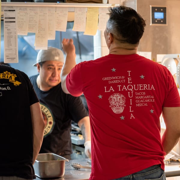 La Taqueria chefs in a busy kitchen, one wearing a red La Taqueria shirt.