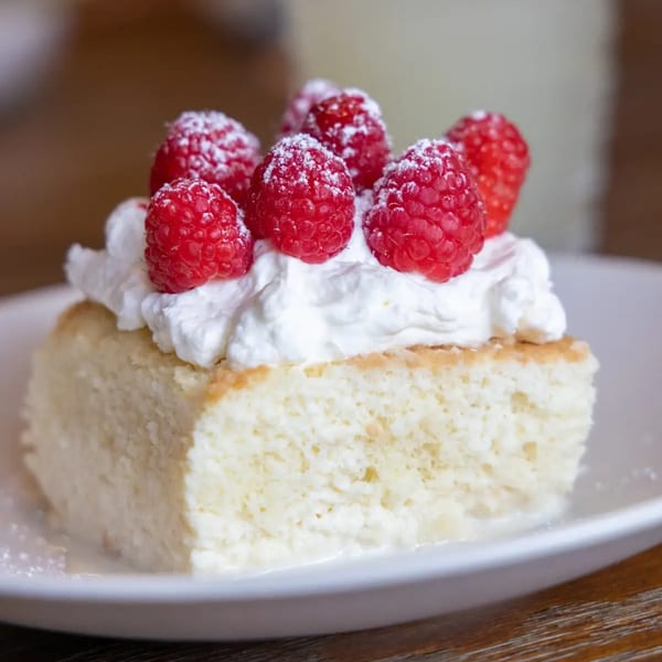 A slice of tres leches cake topped with whipped cream and fresh raspberries on a white plate.