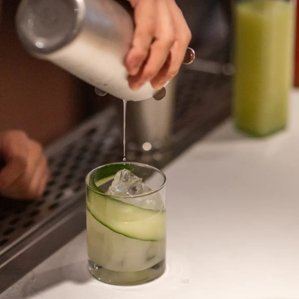 A bartender pours a light green cocktail with cucumber slices and ice into a glass.