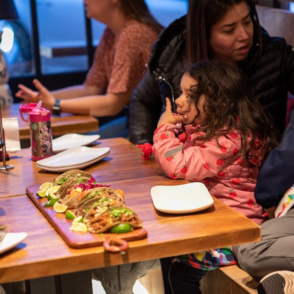 A child in a pink jacket sits at a table with tacos and adults in a La Taqueria restaurant.