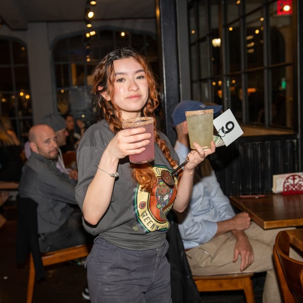 A young woman in a La Taqueria shirt holds two margaritas with patrons in the background.