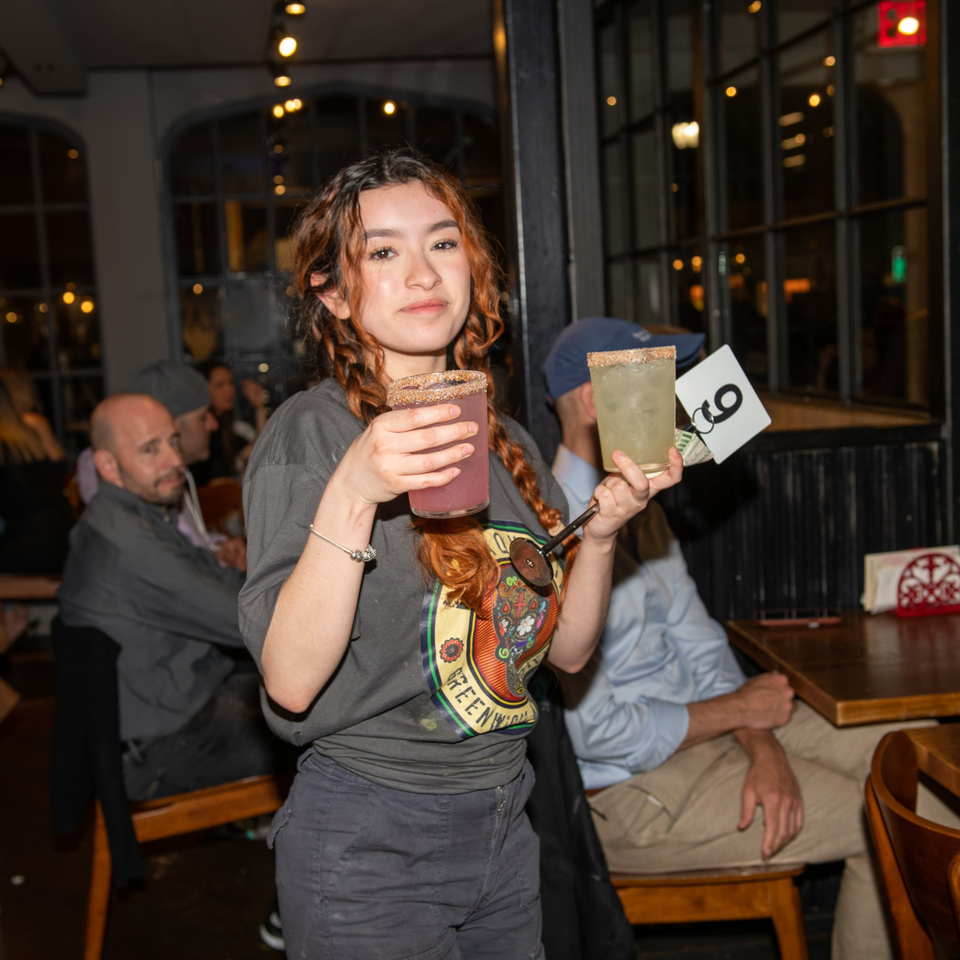 A young woman in a La Taqueria shirt holds two margaritas with patrons in the background.