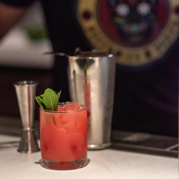 A vibrant red cocktail with mint garnish sits on a bar next to a cocktail shaker.