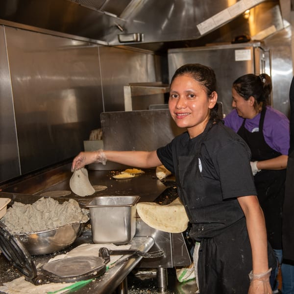 A smiling La Taqueria chef in a black apron prepares tortillas on a griddle in a busy kitchen.