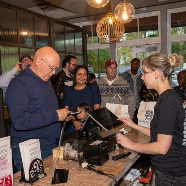 Customers queue at La Taqueria's counter, with a cashier assisting a man paying on his phone.