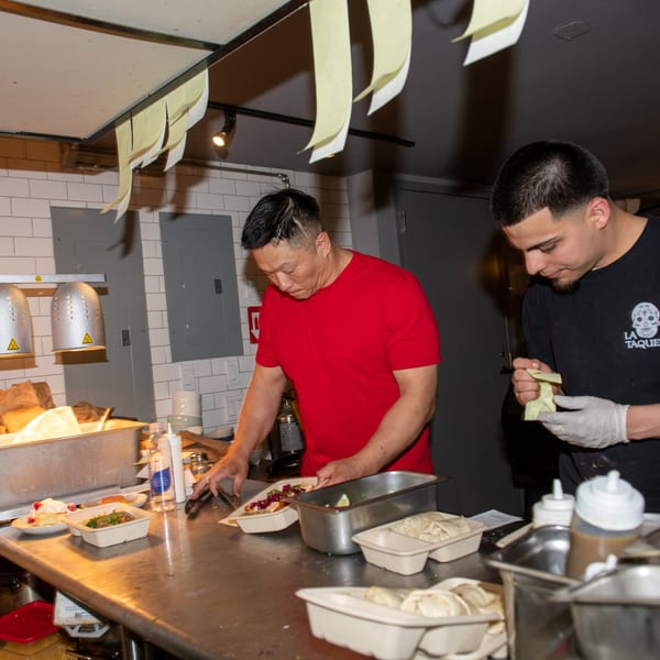 Two La Taqueria chefs preparing Mexican food orders in a busy kitchen.