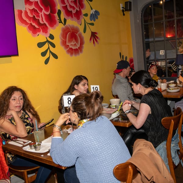 Diners seated at wooden tables in La Taqueria, with a vibrant yellow wall and floral murals.