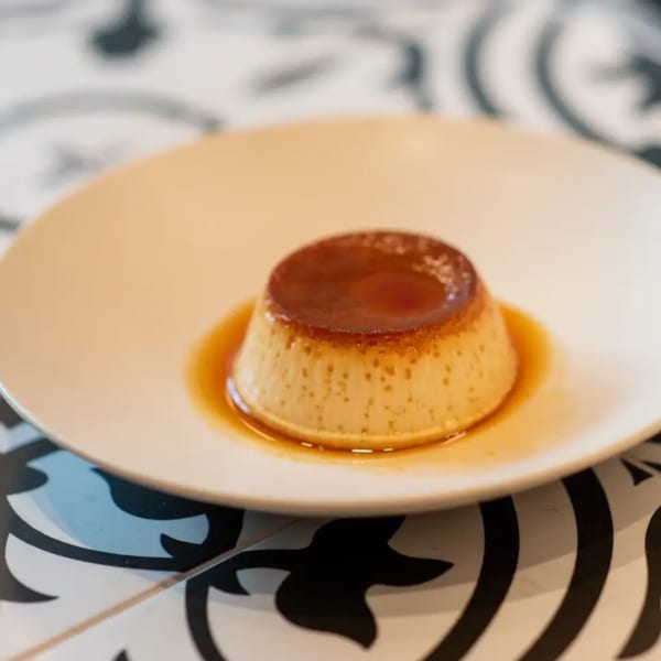 A flan dessert with caramel sauce on a white plate, set on a patterned table.