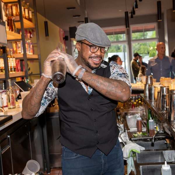 A smiling bartender in a flat cap and glasses shakes a drink at La Taqueria.