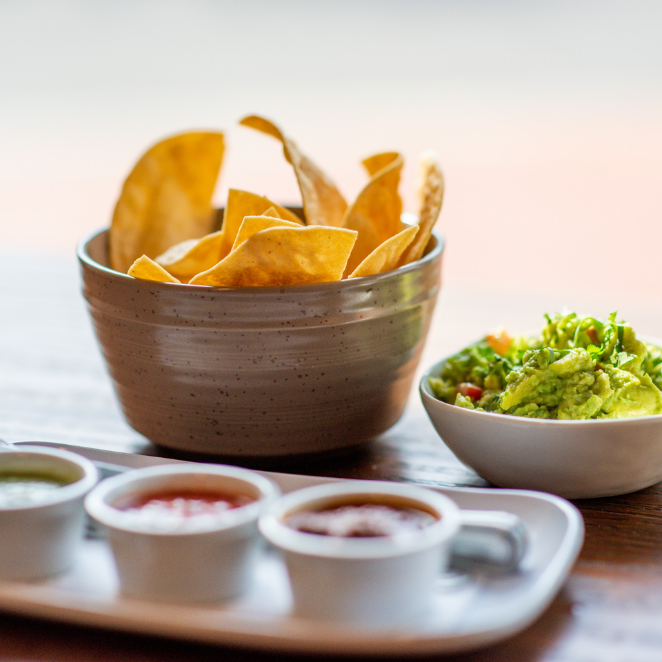 Bowl of tortilla chips with guacamole and assorted dipping sauces on a wooden table.