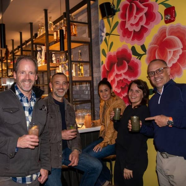 Five smiling people holding drinks at a bar with a vibrant floral mural at La Taqueria.
