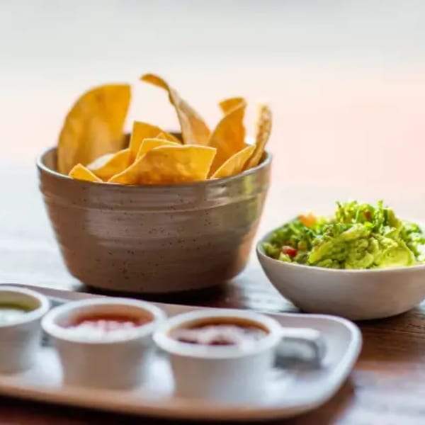 A bowl of tortilla chips, guacamole, and three small dipping sauces on a wooden table.