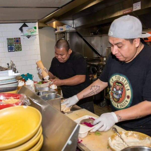 Two La Taqueria chefs in a busy kitchen preparing Mexican food with various ingredients.