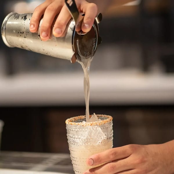 Bartender pouring a margarita from a shaker into a glass with ice and a chili-salt rim.
