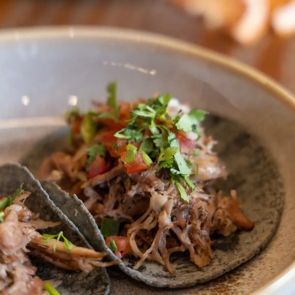 Close-up of two La Taqueria carnitas tacos on blue corn tortillas, topped with pico de gallo.