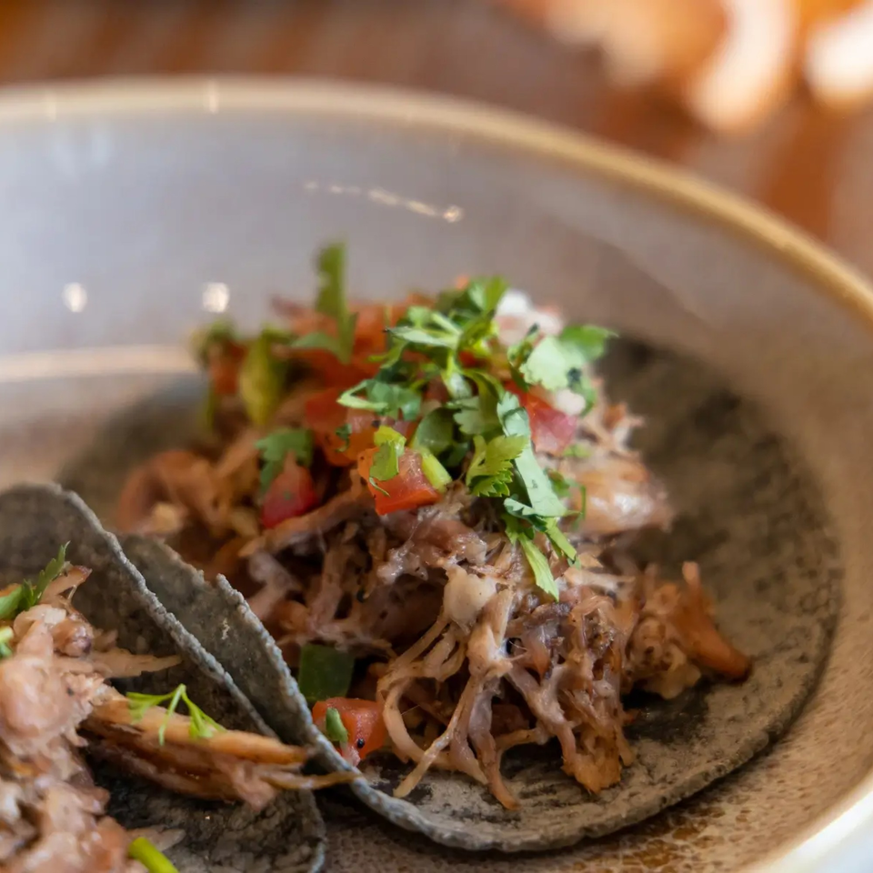 Close-up of two La Taqueria carnitas tacos on blue corn tortillas, topped with pico de gallo.