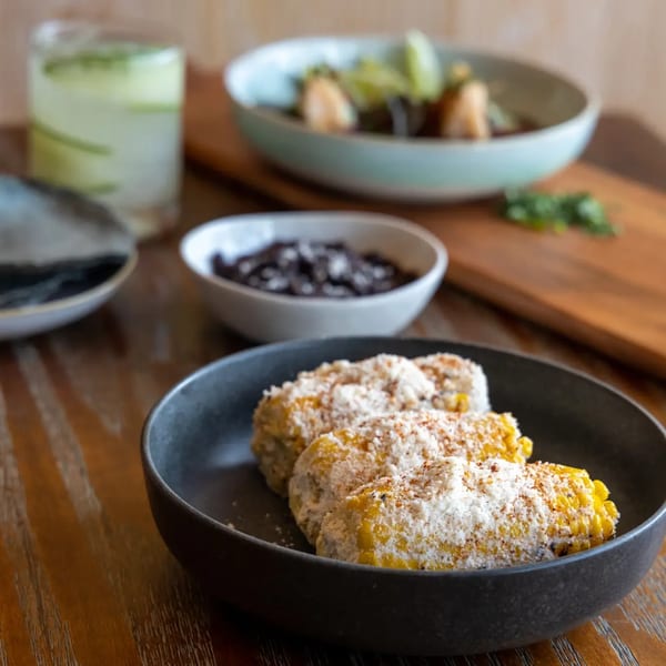 Elote (Mexican street corn) with cotija cheese, black beans, and a drink on a wooden table.