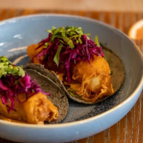 Two La Taqueria fish tacos with red cabbage and cilantro on blue corn tortillas in a bowl.