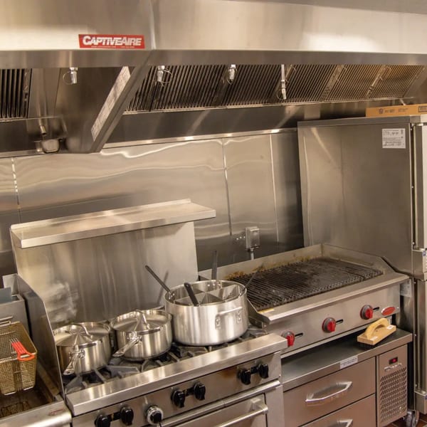A clean, stainless steel commercial kitchen with a grill, stove, pots, and a fryer under a large hood.