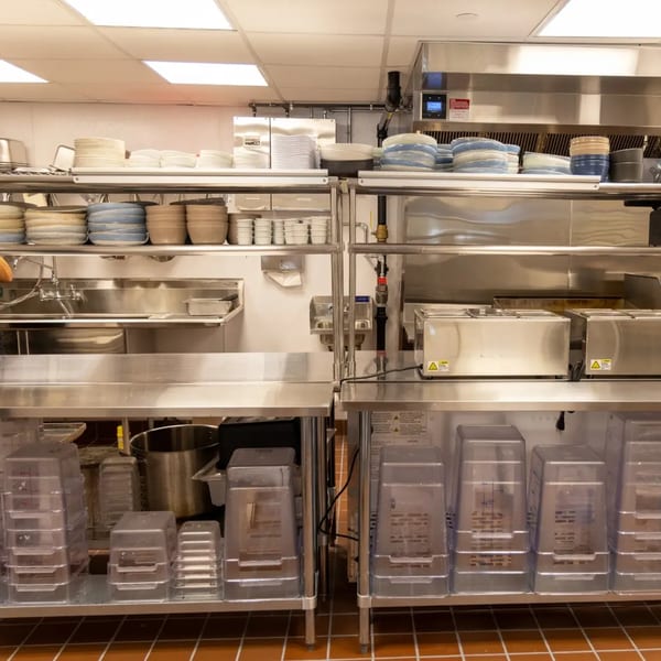 A clean, organized commercial kitchen with stainless steel counters, sinks, and shelves stacked with dishes.
