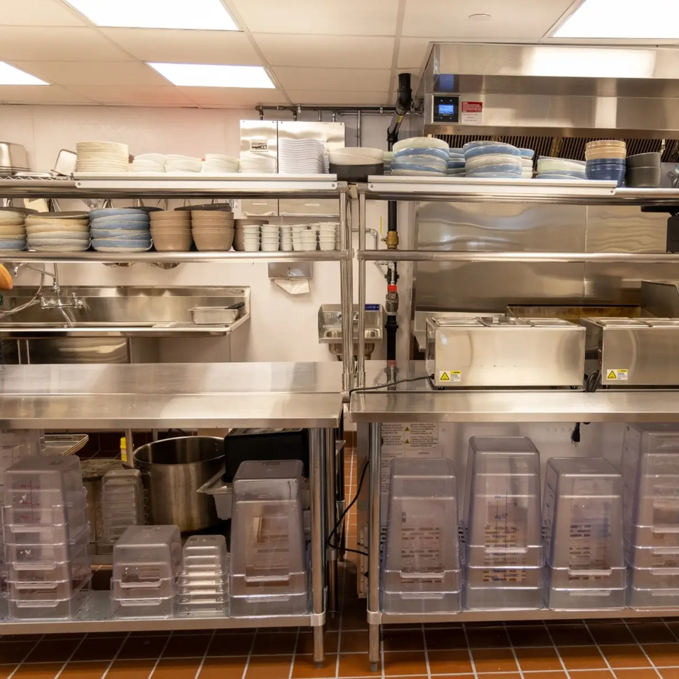 A clean, organized commercial kitchen with stainless steel counters, sinks, and shelves stacked with dishes.